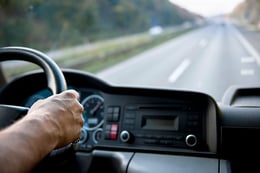 Interior of semi truck driving down the road, looking out windshield and hand on steering wheel.