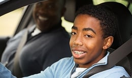 Teen sitting in driver seat of car with father by his side.