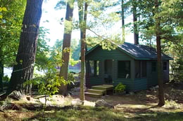A small green cabin with lots of windows surrounded by trees and water.